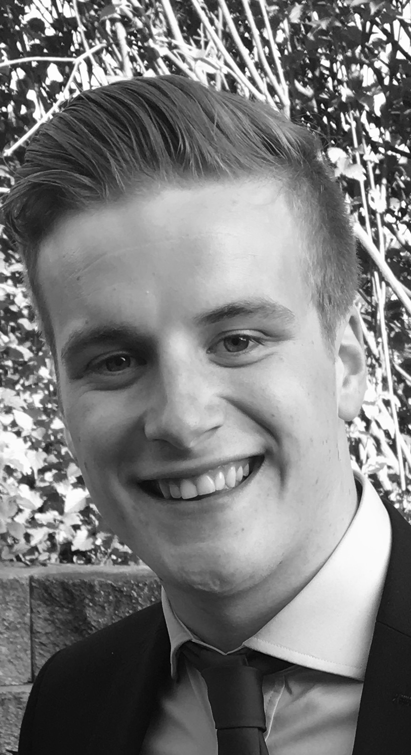 John smiling broadly, in a suit and tie, photographed outdoors against a garden hedge